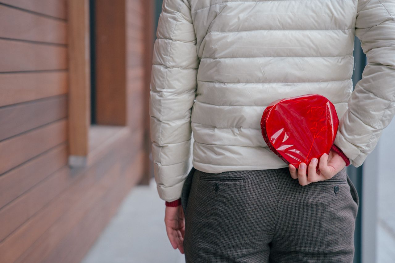 Back view of anonymous male hiding wrapped hear shaped present box while standing on street during holiday celebration in city