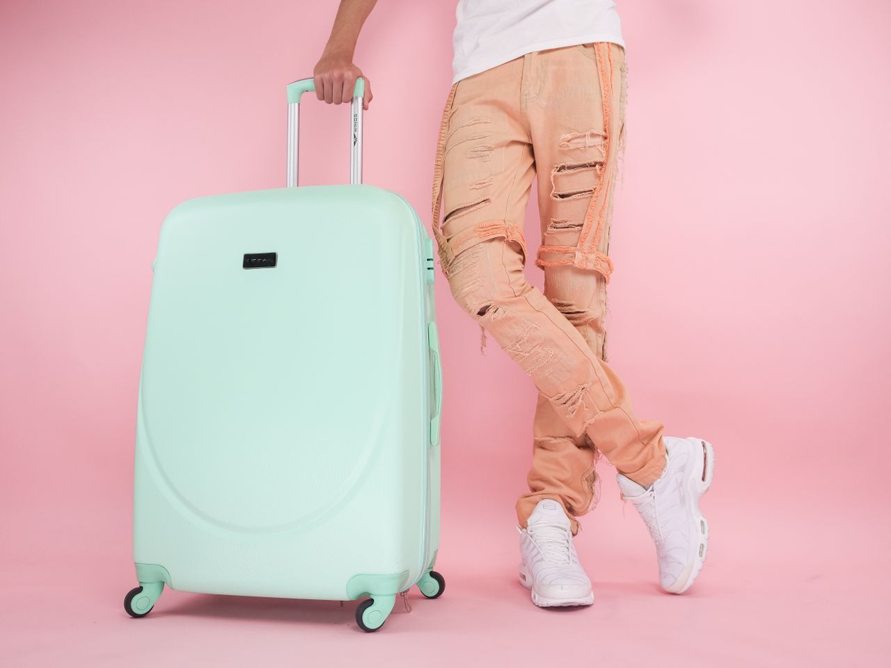 A fashionable studio shot of a person with pastel luggage and trendy ripped jeans against a pink background.